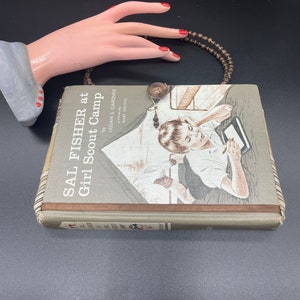 May include: A vintage book titled "Sal Fisher at Girl Scout Camp" by Lillian S. Gardner, illustrated by Marie Stevens. The book cover features a girl sitting on a bed, writing in a notebook. The book is brown with a green spine and a brown leather-like trim. A brown beaded necklace is draped over the book.
