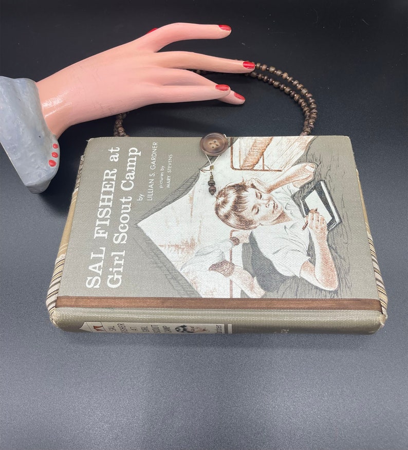May include: A vintage book titled "Sal Fisher at Girl Scout Camp" by Lillian S. Gardner, illustrated by Marie Stevens. The book cover features a girl sitting on a bed, writing in a notebook. The book is brown with a green spine and a brown leather-like trim. A brown beaded necklace is draped over the book.