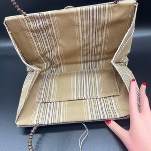 May include: A beige striped fabric clutch bag with a wooden bead handle. The bag has a small pocket on the inside and a white tassel.