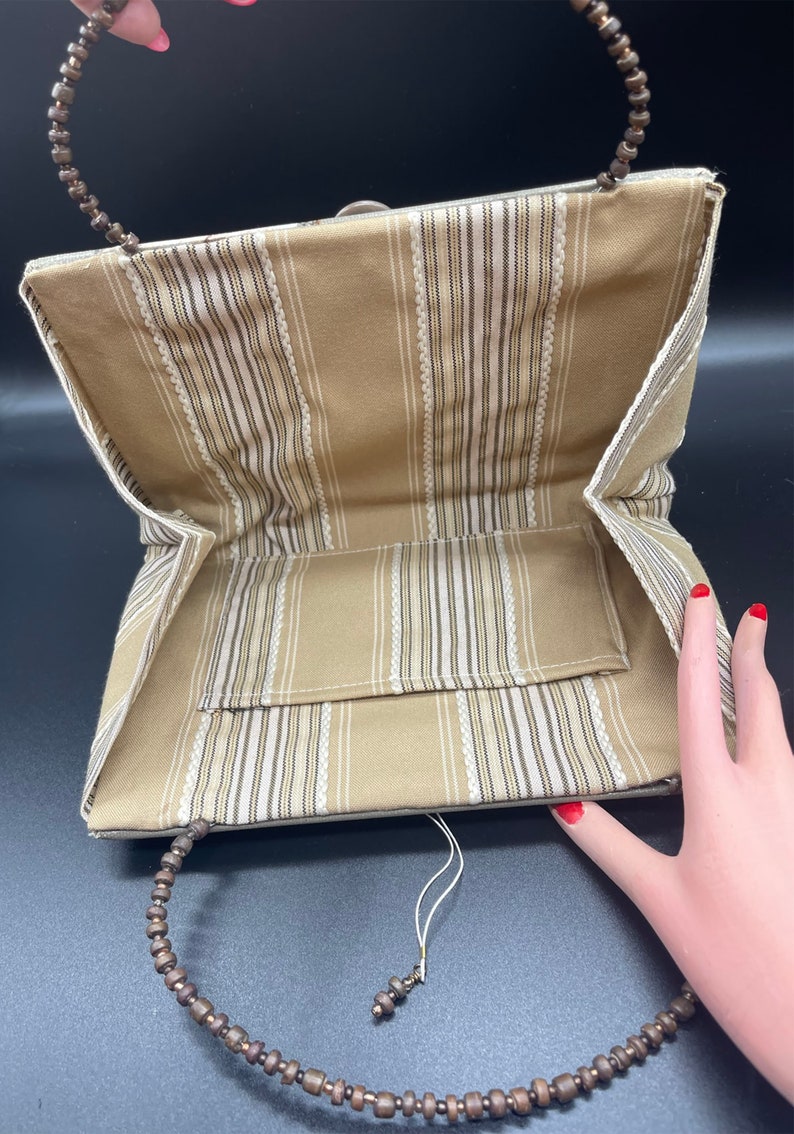 May include: A beige striped fabric clutch bag with a wooden bead handle. The bag has a small pocket on the inside and a white tassel.