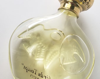 May include: Clear glass perfume bottle with a gold-colored, ornate cap. The bottle has a unique shape with a bird design etched into the glass. The text "L'Air du Temps" and "Nina Ricci" are printed on the bottle.