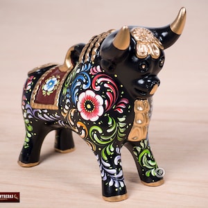 May include: Black ceramic bull figurine with colorful floral designs painted on its body. The bull has gold accents and a saddle.