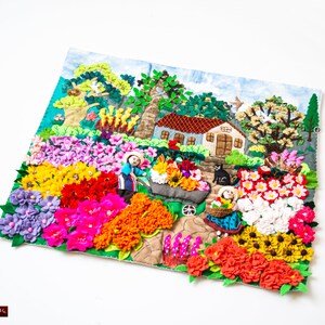 May include: A colourful textile art piece depicting a vibrant garden scene. The artwork features a house, trees, and figures, surrounded by an array of felt flowers in shades of pink, yellow, red, and purple. The piece is set against a light blue background.