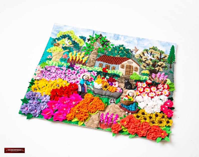 May include: A colourful textile art piece depicting a vibrant garden scene. The artwork features a house, trees, and figures, surrounded by an array of felt flowers in shades of pink, yellow, red, and purple. The piece is set against a light blue background.