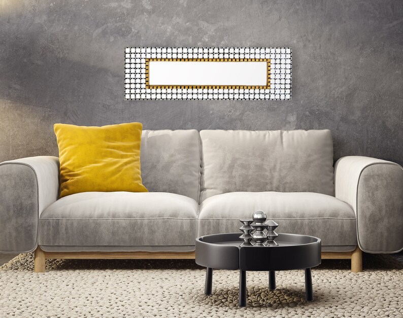 May include: A gray sofa with a yellow pillow sits in front of a gray wall with a rectangular mirror with a gold frame. A black coffee table with a silver sculpture sits in front of the sofa.