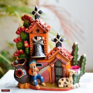 Op de afbeelding: Een kleurrijk keramiek beeldje dat een Mexicaans dorpsscene voorstelt. Het beeldje heeft een kerk met een klokkentoren, een klein huis, een cactus, een lama en een persoon die een muziekinstrument bespeelt. Het beeldje is geschilderd in felle kleuren, waaronder rood, oranje, geel, groen en blauw.