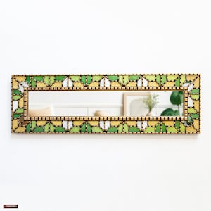 Puede incluir: Un espejo rectangular con un marco dorado y un patrón geométrico de cuadrados verdes, blancos y amarillos. El espejo está colgado en una pared blanca.