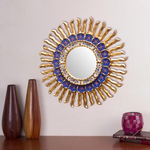 May include: A round mirror with a gold sunburst frame and a blue and white tiled design. The mirror is hanging on a white wall.