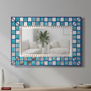 Pode incluir: Espelho retangular com borda de azulejos azuis e brancos. A borda apresenta quadrados alternados de vidro espelhado e azulejos com motivos florais. A moldura interna é prateada e o espelho reflete uma sala com móveis brancos e uma planta.