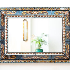 Pode incluir: Espelho de parede retangular com moldura decorativa. A moldura apresenta um fundo azul com padrões florais e de folhas em tons de castanho, amarelo e branco. O espelho reflete uma sala com uma planta e outro espelho.