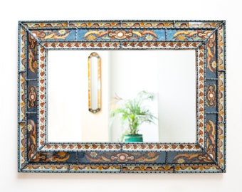 May include: Rectangular wall mirror with a decorative frame. The frame features a blue background with floral and leaf patterns in shades of brown, yellow, and white. The mirror reflects a room with a plant and another mirror.