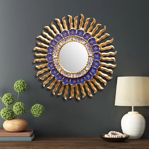 The 17.7 inch Peruvian sunburst mirror hangs above a sideboard in a dining room, where the warm gold carving reads from the far end of the table and the sapphire blue floral ring of reverse painted glass is the detail that rewards a closer look up close.