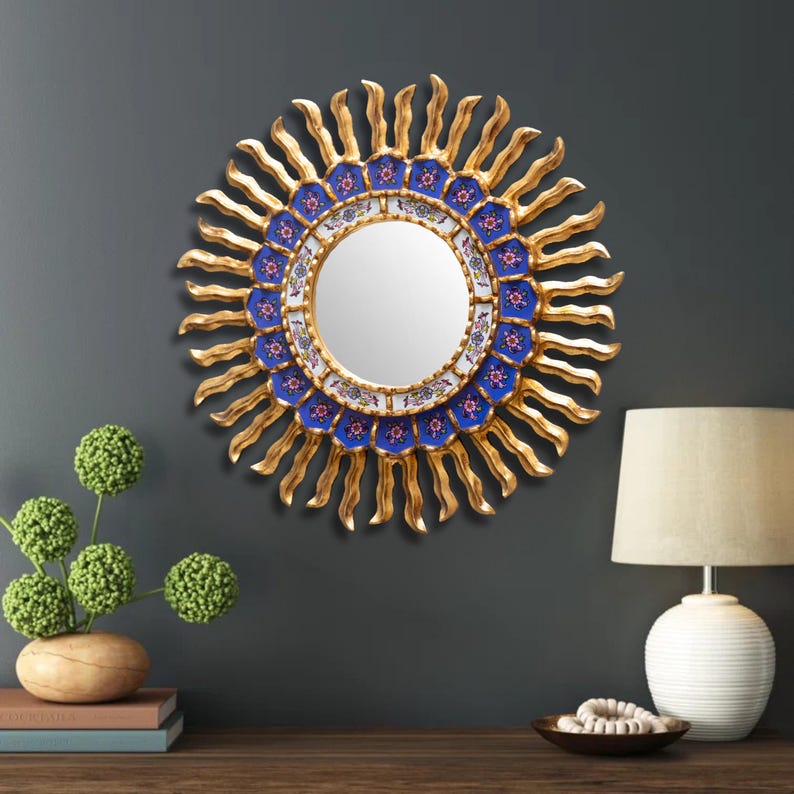 The 17.7 inch Peruvian sunburst mirror hangs above a sideboard in a dining room, where the warm gold carving reads from the far end of the table and the sapphire blue floral ring of reverse painted glass is the detail that rewards a closer look up close.
