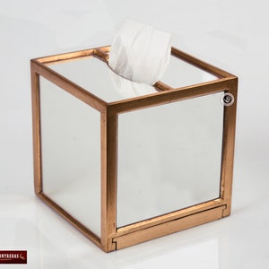 K&ouml;nnte beinhalten: Ein goldfarbener, spiegelnder Taschentuchhalter mit quadratischer Form und einer transparenten Glasoberfl&auml;che. Die Box hat einen goldenen Rand um die Kanten und ein kleines goldenes Etikett an der Seite.