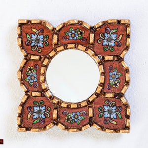 May include: Square decorative mirror with a round center. The frame is brown with gold trim and features floral designs in blue, yellow, and green. The mirror is designed to be hung on a wall.