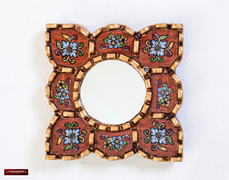 May include: Square decorative mirror with a round center. The frame is brown with gold trim and features floral designs in blue, yellow, and green. The mirror is designed to be hung on a wall.