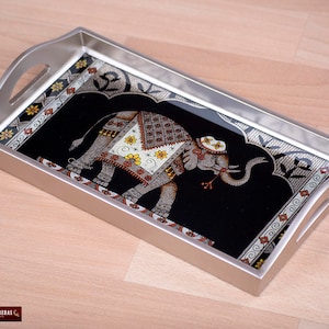 May include: A silver serving tray with a black background and an ornate design featuring an elephant with a floral pattern. The tray has a silver rim and two handles.