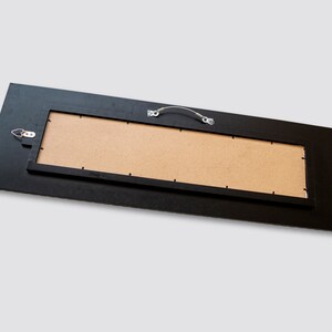 May include: The back of a black framed rectangular mirror with a brown cardboard backing. The mirror has a metal hanging wire and a hook for hanging.