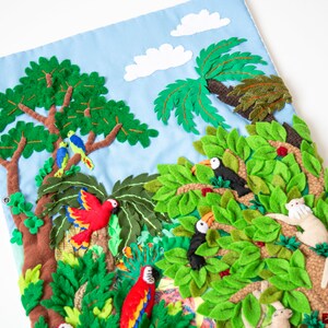May include: A felt wall hanging depicting a jungle scene with toucans, parrots, monkeys, and lush green foliage. The background is a light blue sky with white clouds.