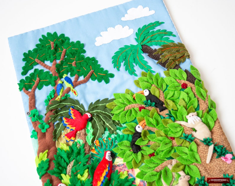 May include: A felt wall hanging depicting a jungle scene with toucans, parrots, monkeys, and lush green foliage. The background is a light blue sky with white clouds.