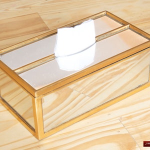 May include: A gold mirrored tissue box holder with a white tissue inside. The box has a rectangular shape with a gold frame.