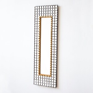 May include: A rectangular mirror with a black and white checkered frame and a gold border. The mirror is hanging on a white wall.