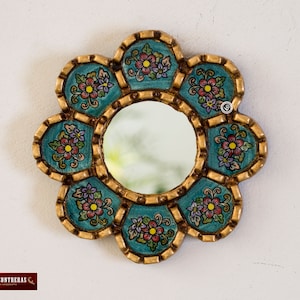Puede incluir: Un peque&ntilde;o espejo redondo con un marco decorativo. El marco est&aacute; hecho de madera y est&aacute; pintado en un color azul verdoso con detalles dorados. El marco est&aacute; decorado con dise&ntilde;os florales en tonos rosa, morado y verde.
