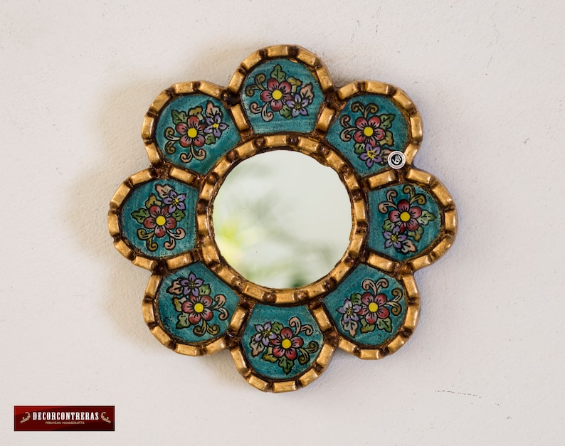 Puede incluir: Un peque&ntilde;o espejo redondo con un marco decorativo. El marco est&aacute; hecho de madera y est&aacute; pintado en un color azul verdoso con detalles dorados. El marco est&aacute; decorado con dise&ntilde;os florales en tonos rosa, morado y verde.