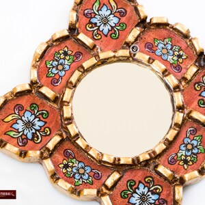 May include: A decorative mirror with a round center and an ornate, multi-lobed frame. The frame is painted in shades of orange and gold, with floral designs in blue, yellow, and green. The mirror is designed to be hung on a wall.