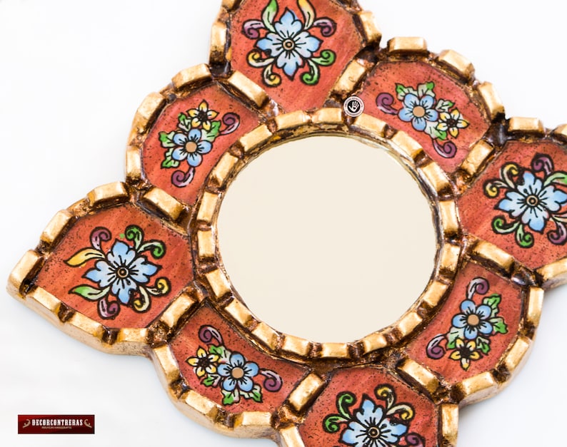 May include: A decorative mirror with a round center and an ornate, multi-lobed frame. The frame is painted in shades of orange and gold, with floral designs in blue, yellow, and green. The mirror is designed to be hung on a wall.
