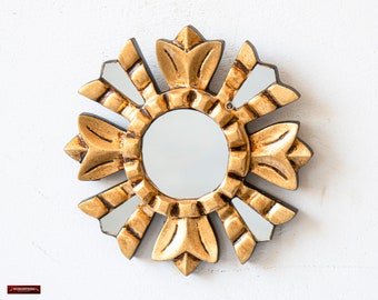 May include: A circular gold-coloured sunburst mirror with a central mirror. The frame has a detailed, carved design with alternating petal-like shapes and rectangular accents. The mirror is mounted on a white wall.