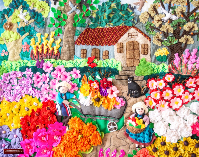 May include: A detailed textile artwork showing a garden scene with a house, trees, flowers, and figures. The piece features a variety of vibrant flowers in shades of pink, red, orange, and yellow. A black cat is near the house.