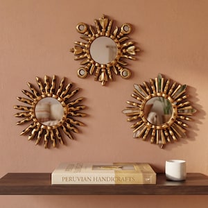 Set of 3 small 6-inch gold sunburst mirrors by UNESCO artisan Edmundo Contreras in an inverted triangle on a terracotta wall. A shelf below with a book shows the scale. Hand-carved mohena wood with bronze leaf. Modern colonial home decor for gallery walls and artisanal accents.