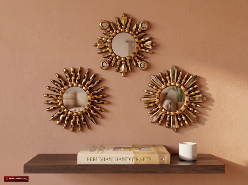 Set of 3 small 6-inch gold sunburst mirrors by UNESCO artisan Edmundo Contreras in an inverted triangle on a terracotta wall. A shelf below with a book shows the scale. Hand-carved mohena wood with bronze leaf. Modern colonial home decor for gallery walls and artisanal accents.