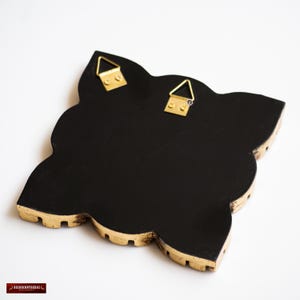 May include: Black decorative wall art with a quatrefoil shape and gold trim. The back of the art piece features two gold-colored metal hangers for easy wall mounting. The edges have a scalloped design.