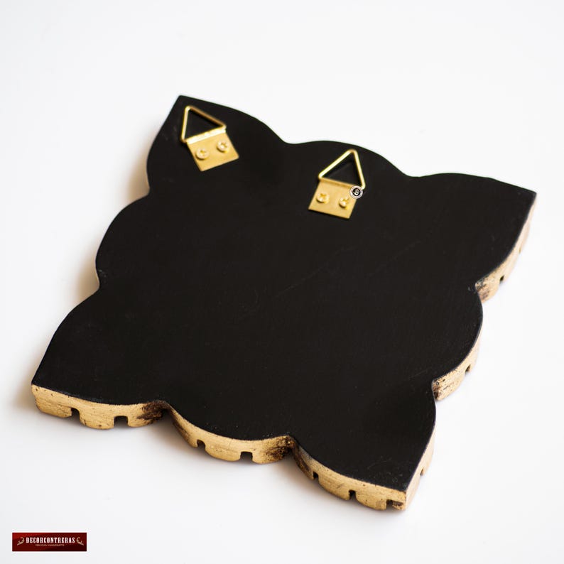 May include: Black decorative wall art with a quatrefoil shape and gold trim. The back of the art piece features two gold-colored metal hangers for easy wall mounting. The edges have a scalloped design.