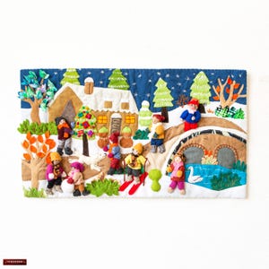 Puede incluir: Un tapiz de pared de fieltro que representa una escena de invierno con una pequeña casa, un puente, un río y varias personas. Las personas llevan ropa de invierno y se dedican a diversas actividades, como esquiar, jugar en la nieve y caminar. La escena se desarrolla sobre un fondo azul con copos de nieve blancos.