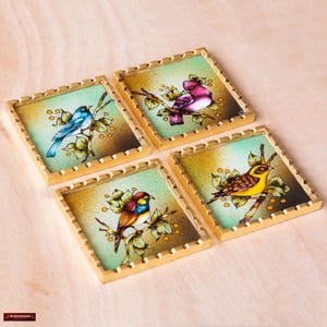 May include: Four square coasters with gold trim and a green background. Each coaster features a different bird with colorful painted details. The birds are a blue bird, a pink bird, a yellow bird, and a red and yellow bird.