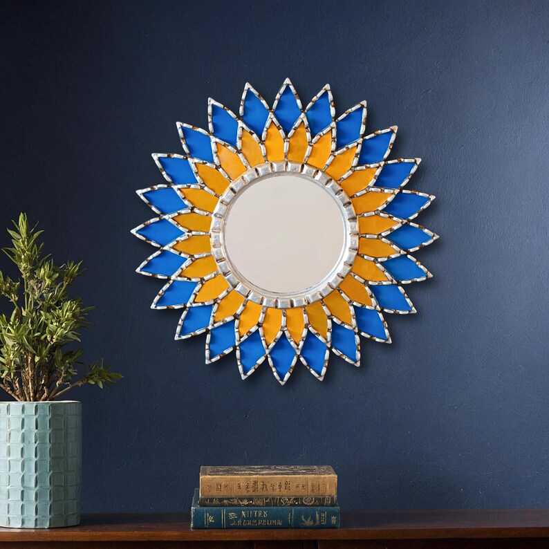 K&ouml;nnte beinhalten: Ein runder Wandspiegel mit einem dekorativen Rahmen im Sonnenblumen-Design. Der Rahmen hat blaue und gelbe rautenf&ouml;rmige Glasteile mit silberfarbenem Metallbesatz. Der Spiegel ist an einer dunkelblauen Wand befestigt.
