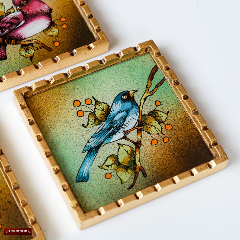 May include: A blue bird with a yellow beak and black eyes sits on a branch with green leaves and orange berries. The bird is perched on a gold-framed coaster with a green and brown background.