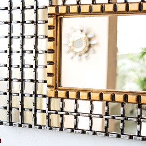 May include: A gold framed mirror with a grid pattern. The mirror is reflecting a room with a plant in the background.