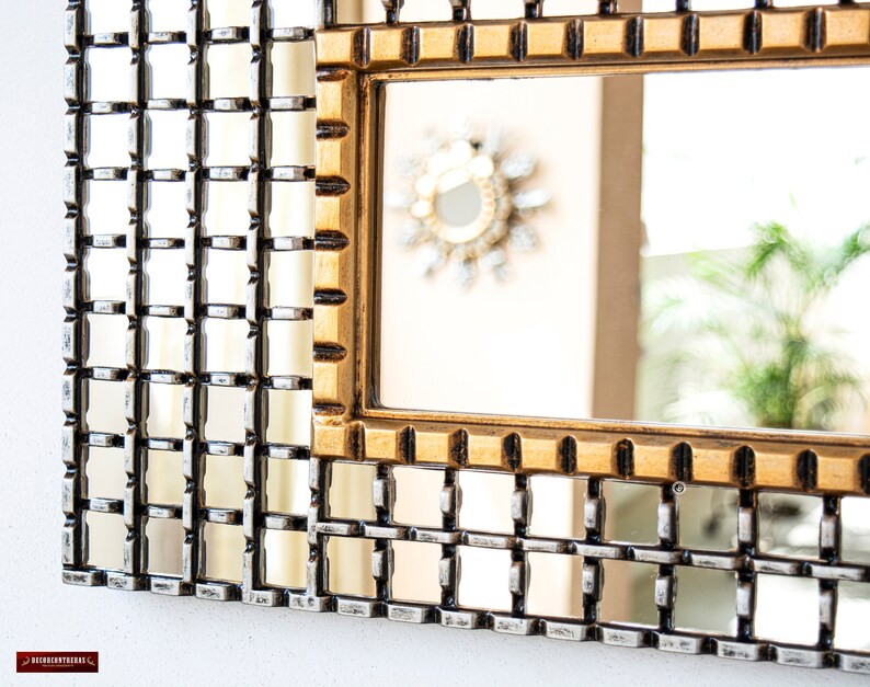 May include: A gold framed mirror with a grid pattern. The mirror is reflecting a room with a plant in the background.