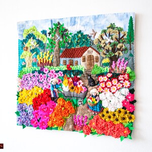 May include: A detailed textile artwork showing a garden scene with a house, trees, and figures. The piece uses a variety of colours, including red, pink, yellow, and green, and textures to depict flowers and foliage. The artwork is mounted on a white wall.