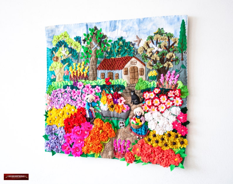 May include: A detailed textile artwork showing a garden scene with a house, trees, and figures. The piece uses a variety of colours, including red, pink, yellow, and green, and textures to depict flowers and foliage. The artwork is mounted on a white wall.