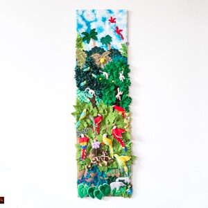 May include: A colorful textile art piece depicting a jungle scene. The artwork features parrots, monkeys, and other animals amidst lush green foliage, trees, and a blue sky with clouds. The piece is rectangular and mounted on a white wall.