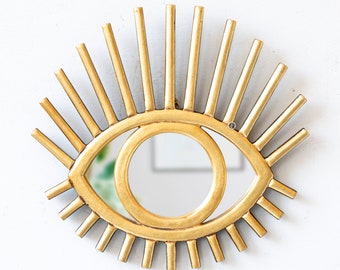 Könnte beinhalten: Ein goldfarbener, augenförmiger Wandspiegel. Der Spiegel hat eine kreisförmige Mitte und ist von einem Rahmen mit radialen Linien umgeben, die Wimpern oder Sonnenstrahlen ähneln. Der Spiegel ist an einer weißen Wand befestigt.
