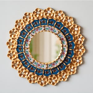 May include: Round decorative mirror with a gold-colored floral frame. The mirror features a blue and white patterned border with floral accents. The central mirror is circular, reflecting the surrounding environment. A stylish home decor item.