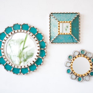 May include: Three decorative mirrors with teal and silver accents are displayed against a white wall. The mirrors have unique shapes: a scalloped circle, a square, and a small circle with a gold and teal border. The mirrors are designed for wall decor.