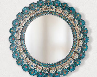 Op de afbeelding: Een ronde wandspiegel met een decoratieve lijst met een geschulpte rand. De lijst is versierd met ingewikkelde bloemmotieven in tinten turquoise, crème, rood en blauw, gerangschikt in herhalende segmenten rond het centrale glazen oppervlak.