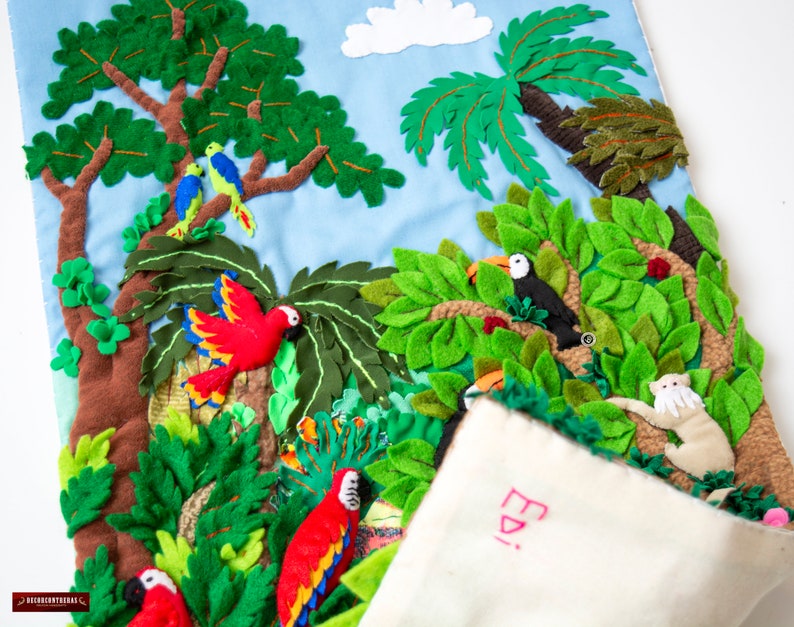 May include: A felt wall hanging depicting a jungle scene with green leaves, brown trees, red and green parrots, a toucan, and a white monkey. The hanging has a white fabric pocket with the word "Edi" written in pink.